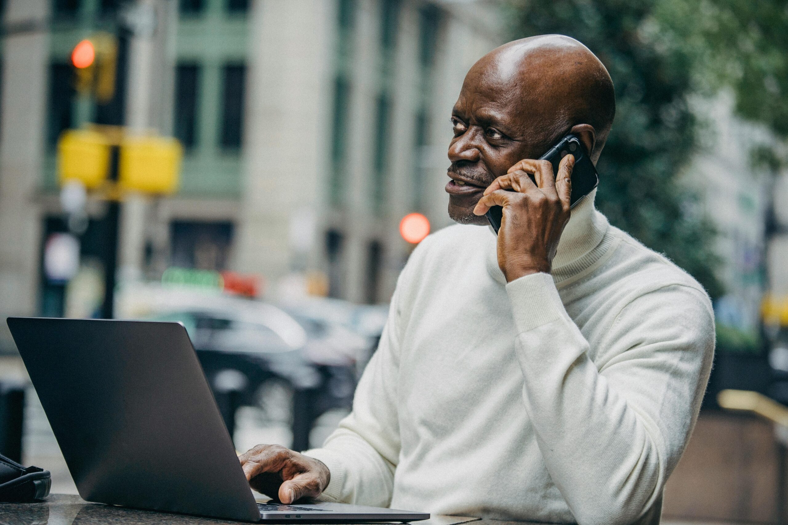 African American man in casual clothes sitting at table with laptop and talking on smartphone during work